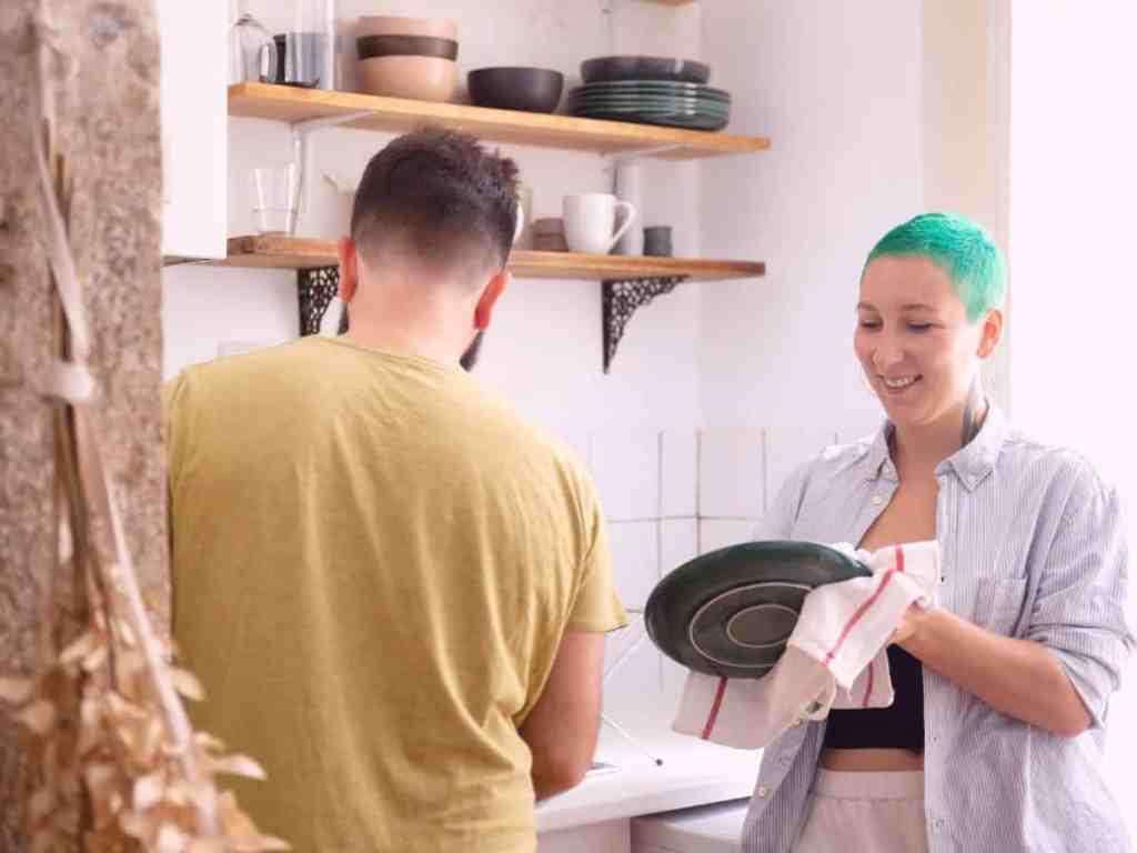 Casal lavando os pratos juntos. Ele, que usa um camiseta bege, está lavando a louça, enquanto ela, de cabelo verde curto e camisa azul clara aberta, seca os pratos.
