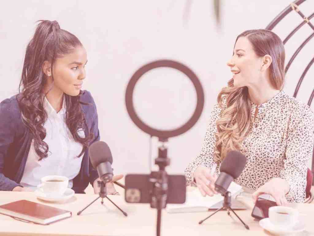 Duas mulheres gravando um podcast. A da esquerda usa camisa branca por dentro e terninho azul royal. Enquanto, a da direita usa uma blusa de manga longa com estampa de animal print.