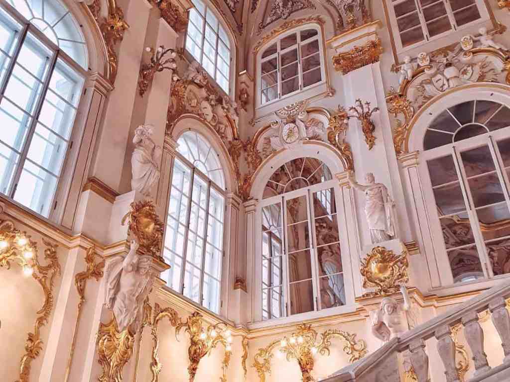 Interior de um prédio ou museu de paredes brancas com ornamentos dourados por toda ela.