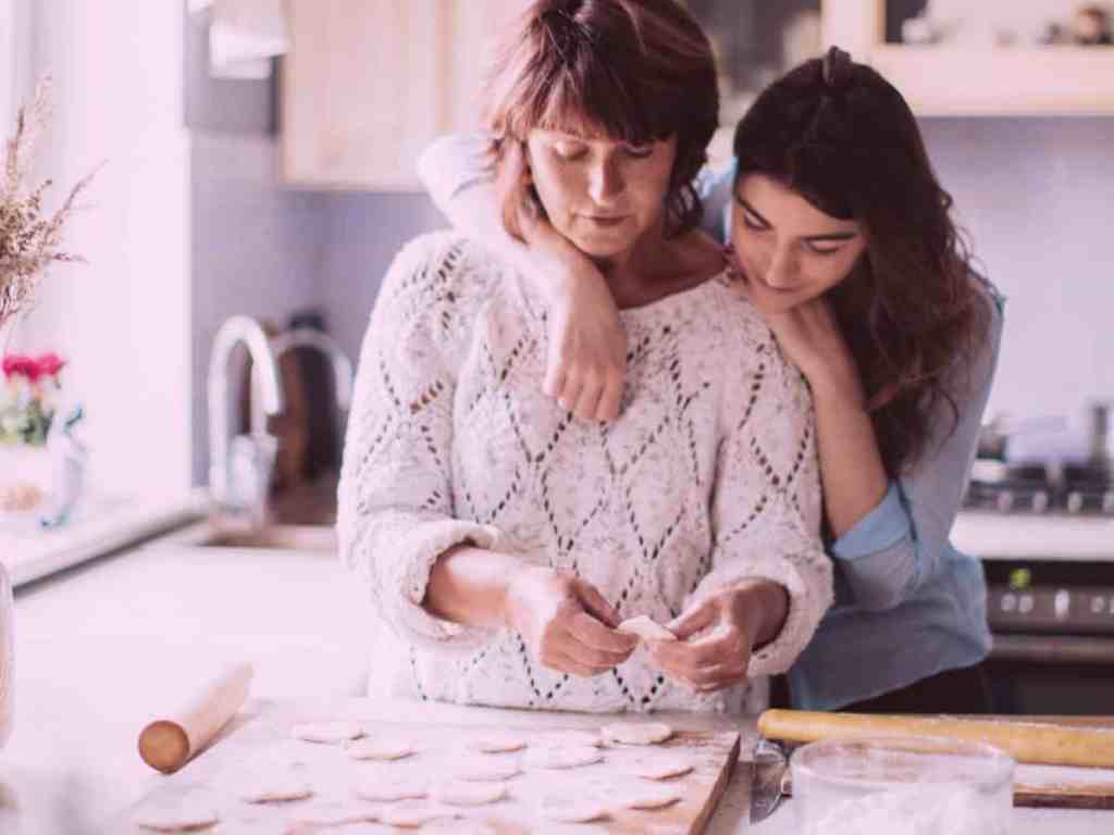 Como lidar com: sair da casa dos pais para se casar - Mae-e-filha-cozinhando-juntas-abraco Filha abraçando a mãe por trás, enquanto a mãe prepara a massa da comida na bancada da cozinha.