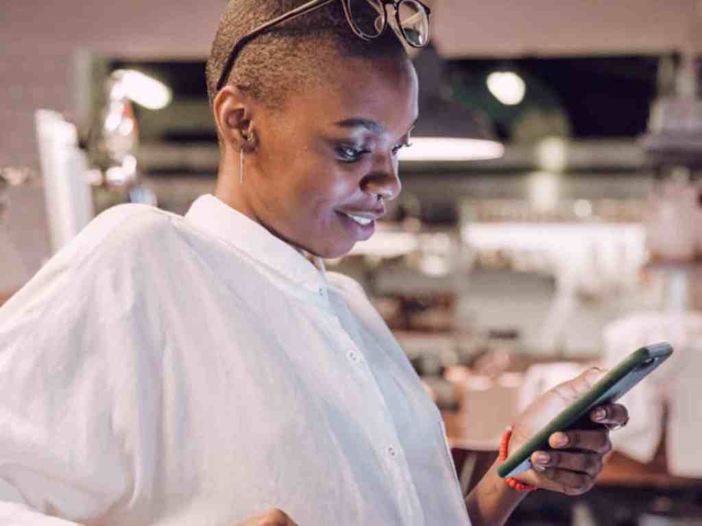 Por que é importante ter um tempo só para você no relacionamento? - Mulher-feliz-mexendo-no-celular Mulher sorrindo enquanto mexe no celular. Ela é careca e está com um óculos de grau na cabeça, usa camisa branca, pulsiera vermelha.