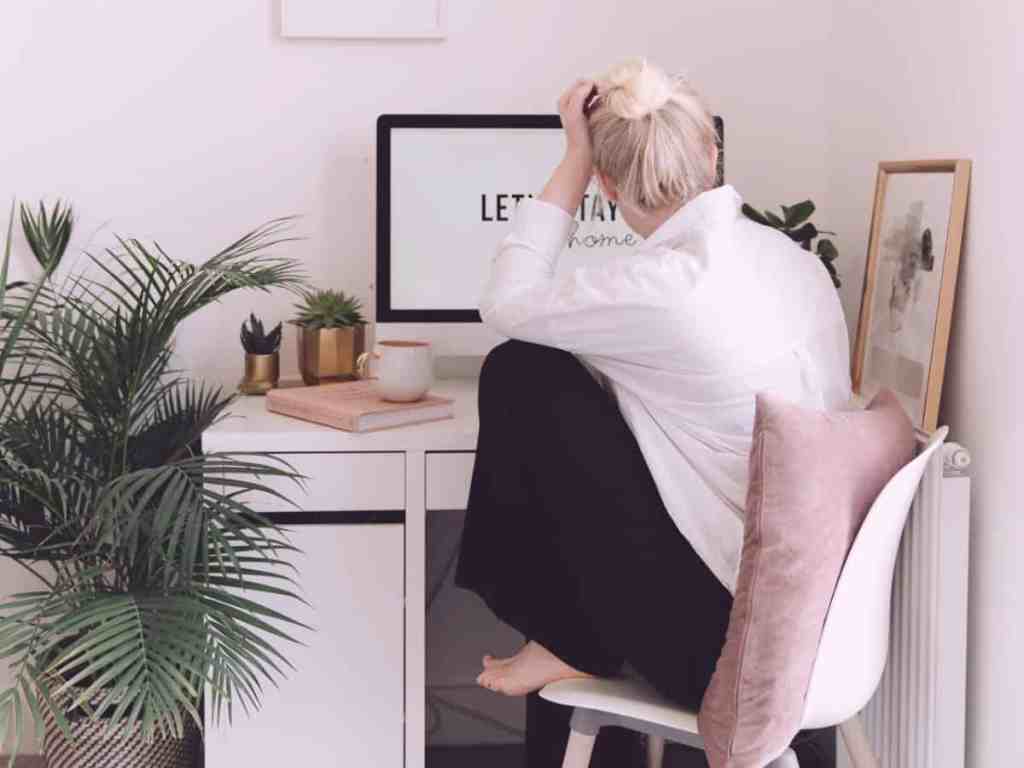 Mulher sentada trabalhando no computador de home office. Ela tem cabelo loiro preso em um coque e usa camisa branca e calça preta.