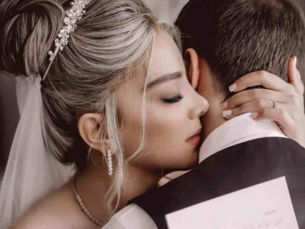 Maquiagem para noiva: 5 opções para arrasar - Noiva-e-noivo-abraco-votos Noivos abraçados. Ela está com o cabelo loiro em coque com tiara e véu. Enquanto segura um papel. Já ele está de costas, sendo abraçado, cabelo castanho curto, camisa branca e colete preto.