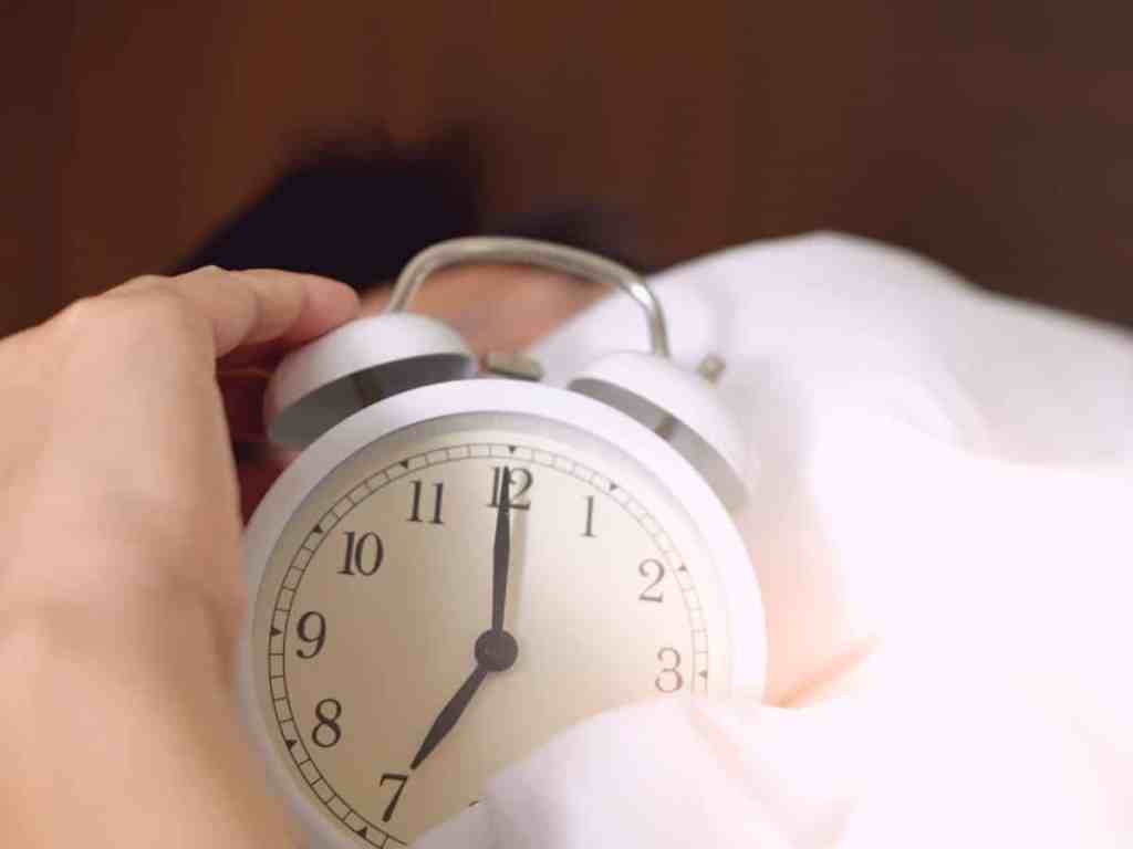 Mão segurando um relógio despertador branco, enquanto um homem está dormindo.