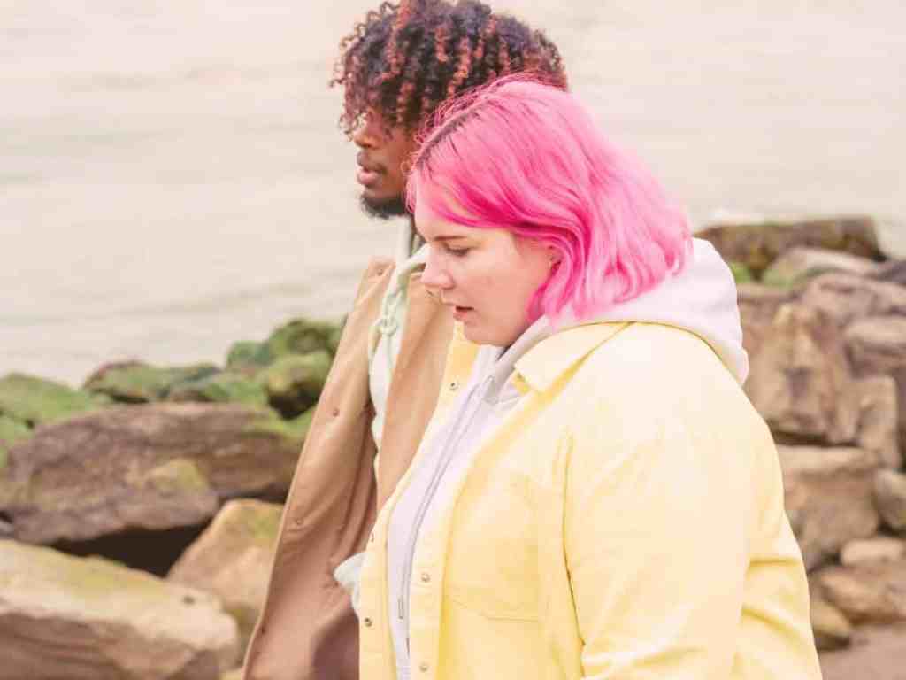Casal andando pelas pedras em frente ao rio. Ela tem cabelo rosa e usa camisa amarela com moletom branco por baixo e ele tem cabelo cacheado e usa sobretudo bege.
