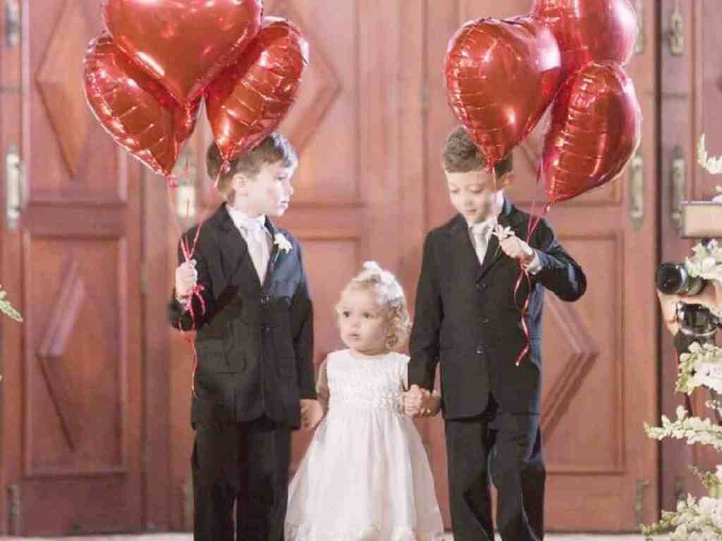 Daminha de honra no meio e dois pajens um de cada lado segurando a mão dela e balões vermelhos em formato de coração.