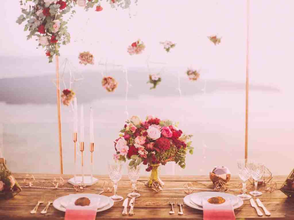 Mesa do casamento com flores decorativas penduradas. Tem buquê no centro, e vários pratos, talheres, taças e um candelabro.