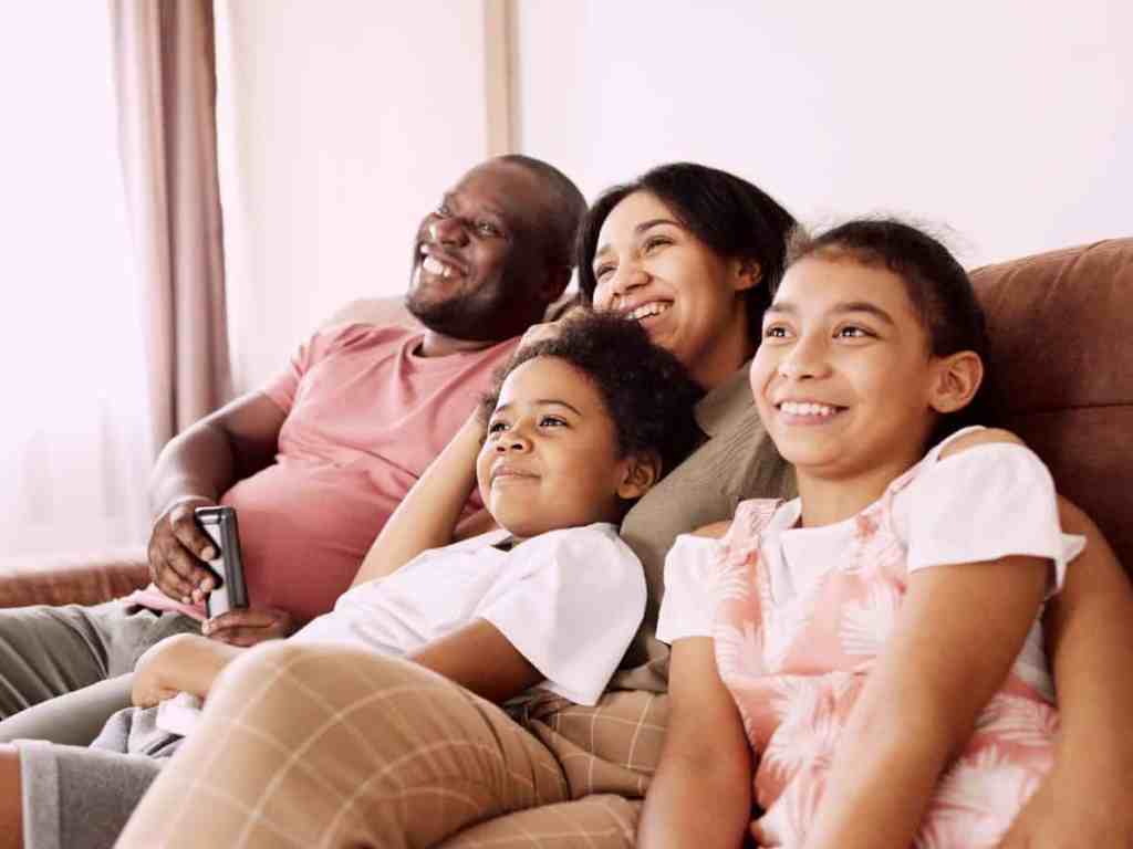 Família sentada no sofá assistindo tv. Tem o pai, a mãe, um filho e uma filha.