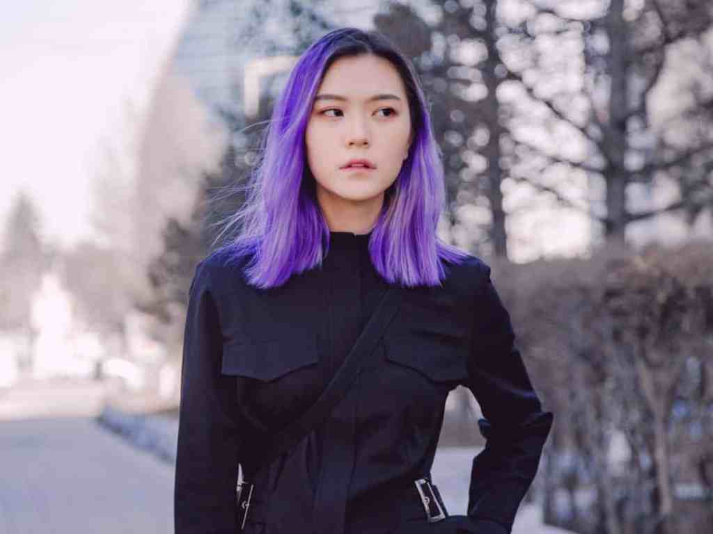 Garota posando para foto. Ela tem cabelo violeta e usa blusa de manga longa preta com bolsos.