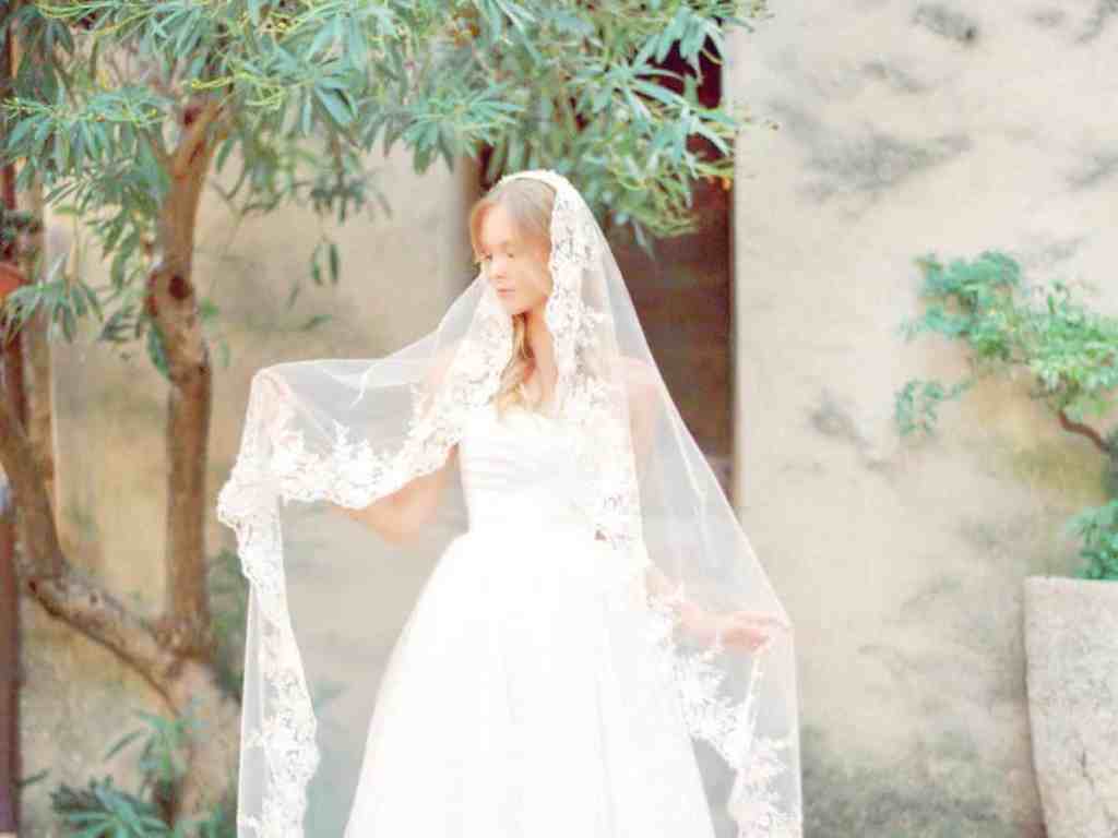 O que é uma mantilha? - Mantilha-noiva Noiva loira usando véu longo segurando-o.