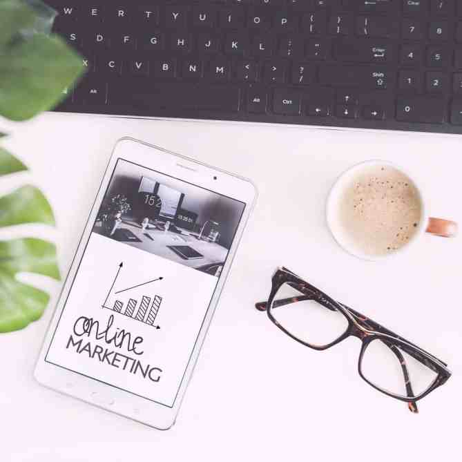 Imagem de um celular em cima de uma escrivaninha, perto do teclado, do óculos de grau e do café. No celular está um desenho de gráfico e embaixo está escrito "online marketing".