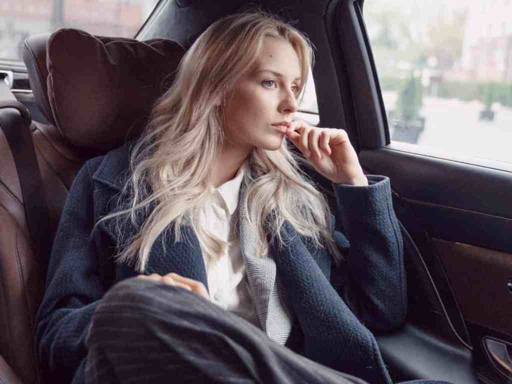 Mulher com terno azul sentada no banco de trás no carro olhando pela janela.