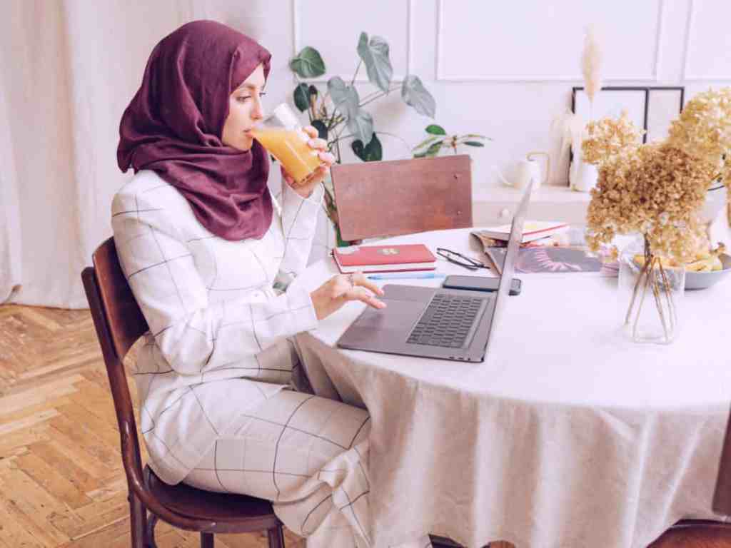 Mulher sentada bebendo suco e mexendo no notebook. Ela usa hijab mársala e conunto de roupas xadrez branco