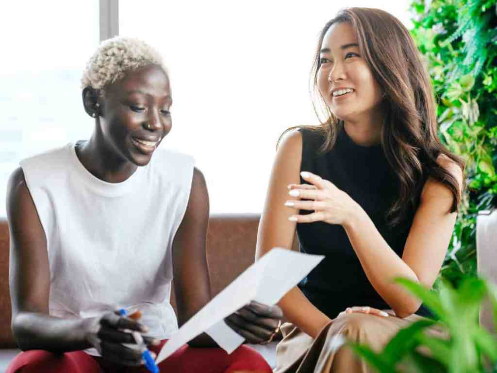 O que é empreendedorismo feminino? - Mulheres-empresarias-projeto Duas mulheres rindo juntas. Uma está olhando para um papel, a outra está olhando para outra pessoa e falando.