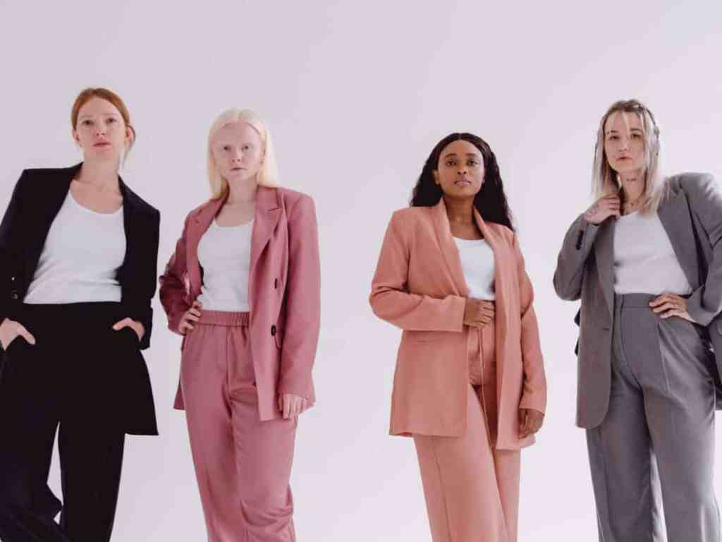 Quatro mulheres olhando para frente, todas estão vestidas com roupa social, terno e calça, mas cada uma de uma cor diferente. Respectivamente os conjuntos são: preto, rosa, coral e cinza.