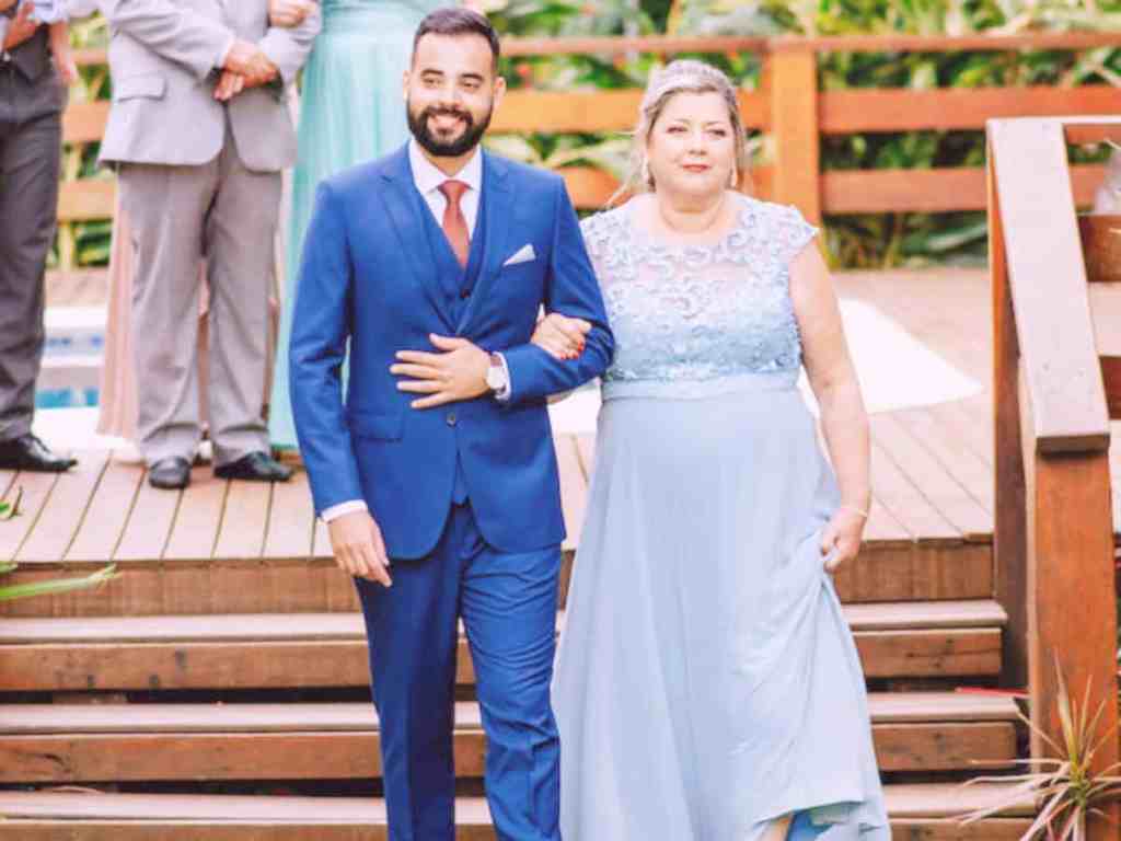 Noivo entrando no casamento acompanhado de sua mãe. Ele de terno azul caneta e gravata vermelha e ela de vestido azul claro.