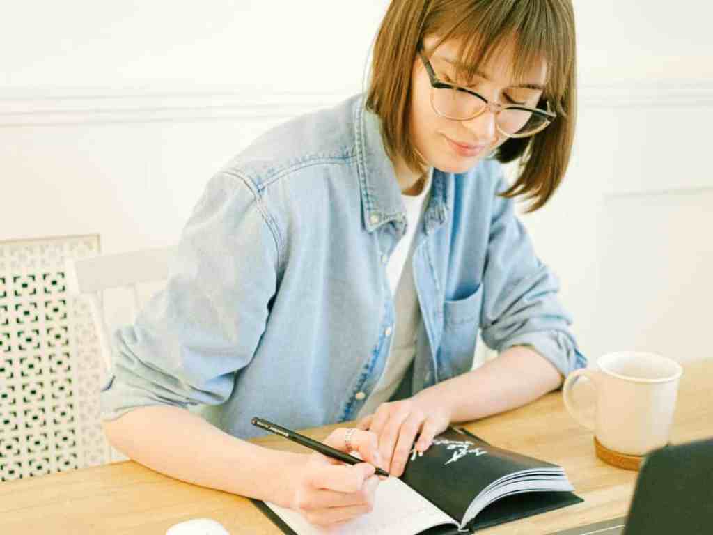 Mulher anotando em um caderno. Ela está usando jaqueta jeans e óculos de grau.