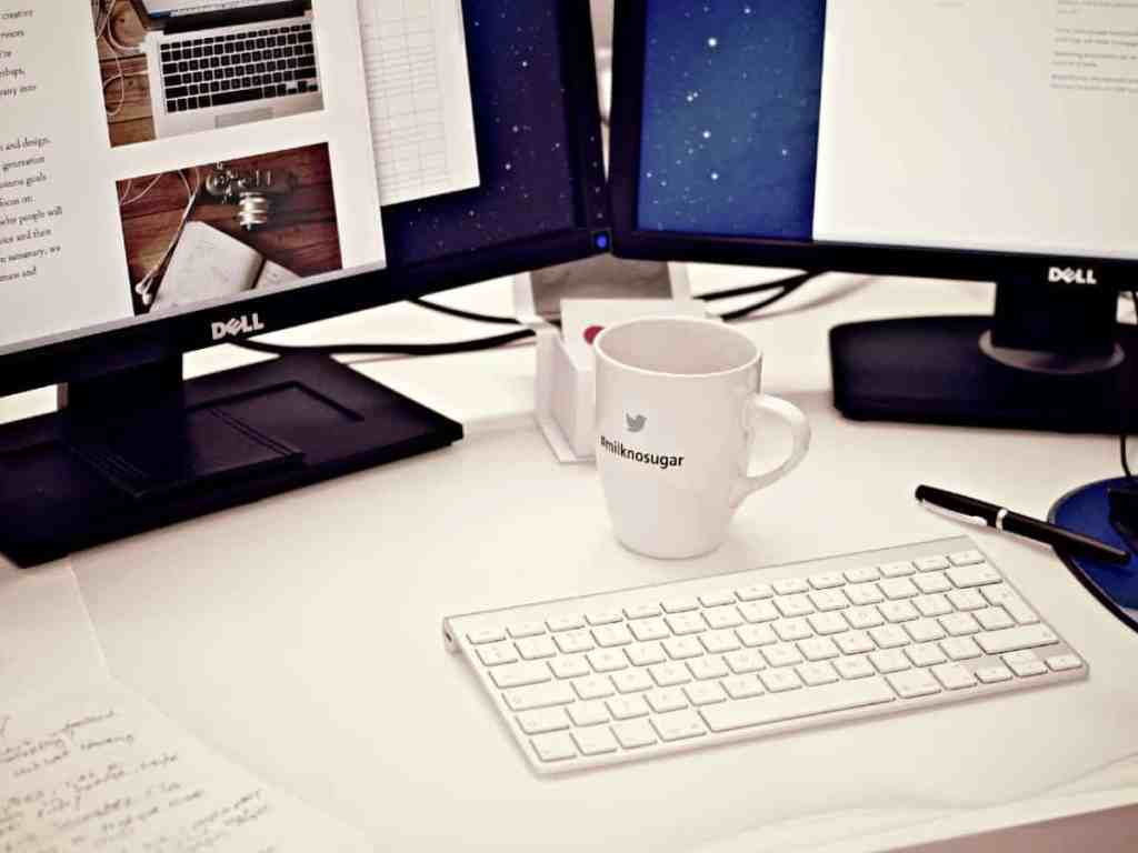 Mesa com computador de duas telas, uma xícara branca e uma folha.