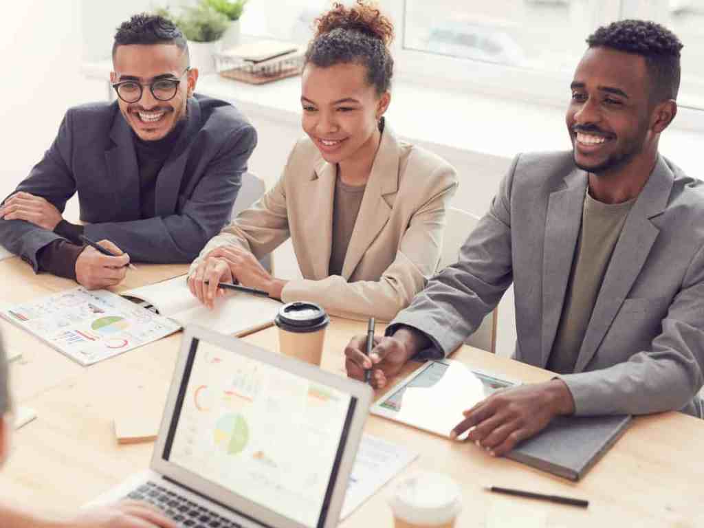 Três pessoas sentadas em uma mesa de reunião, dois homens e uma mulher. Em frente a eles tem alguém com o notebook aberto.