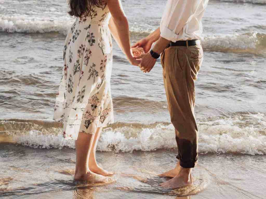 Casal na praia de mãos dadas com pés na água.