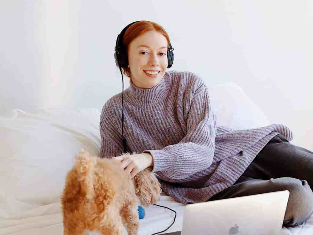 Garota ruiva sorrindo sentada na cama ouvindo música enquanto brinca com seu pet.