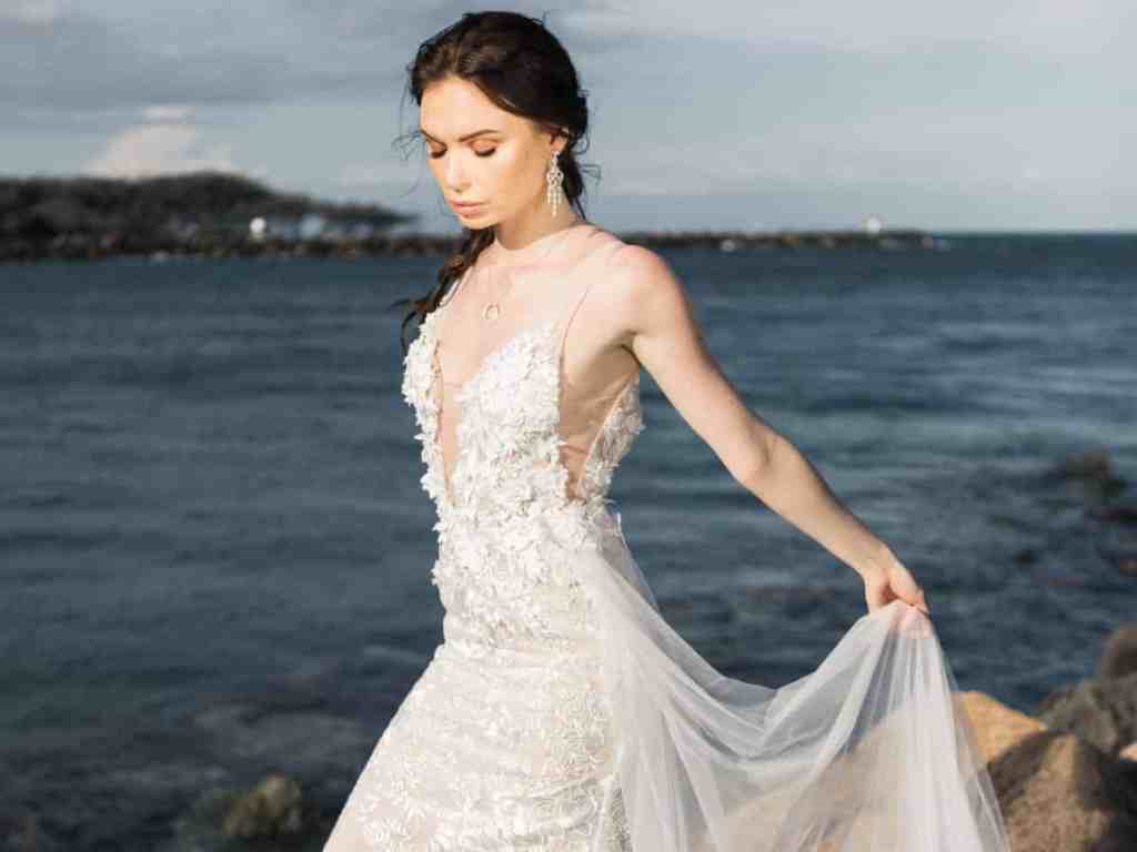 Mulher na praia vestida de noiva segurando o tulê do vestido