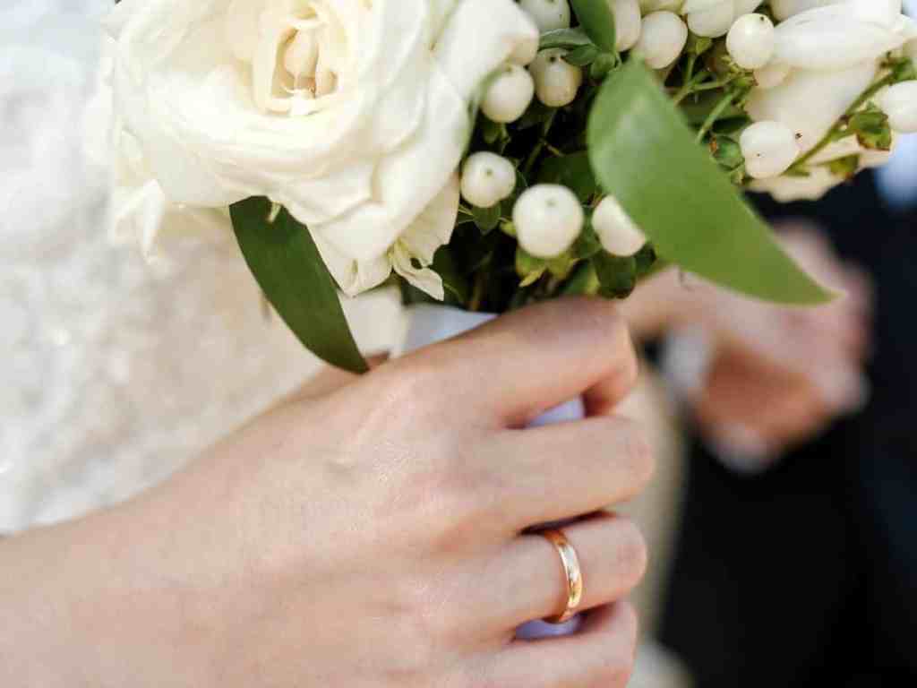 Noiva com aliança rosé no dedo segurando buquê de flores brancas.