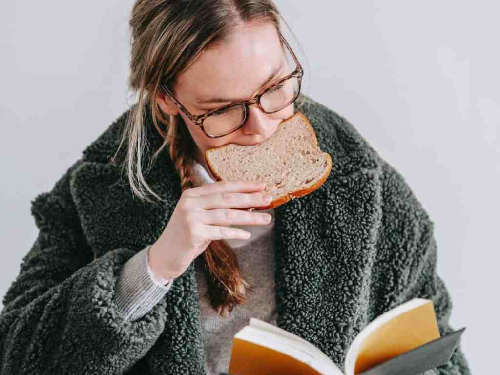 Mulher loira comendo pão de forma enquanto lê