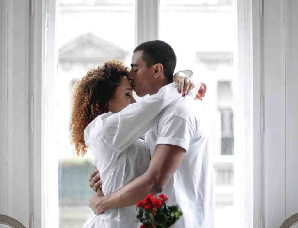 Casal abraçado, ele está dando um beijo na testa dela. Ambos vestem camisas brancaas.