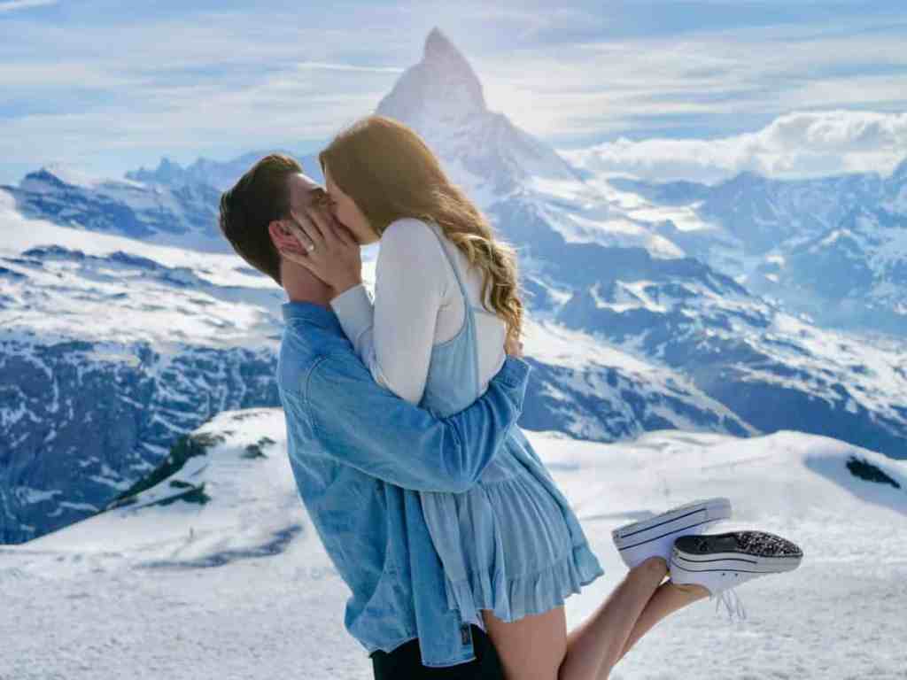 Casal se beijando após pedido de casamento. Fundo da imagem com montanhas