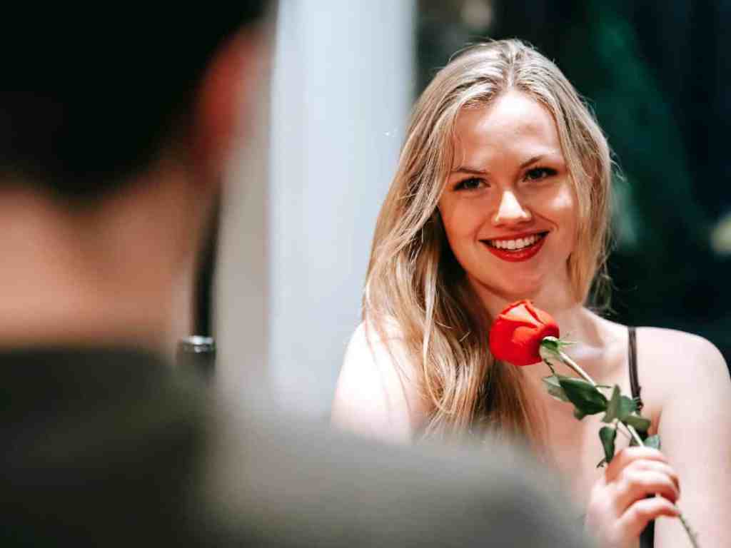 Casal em um encontro. Mulher loira sorri segurando uma rosa vermelha.