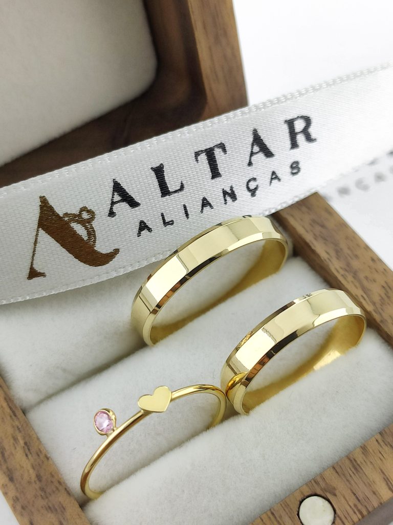 Imagem de caixinha marrom com par de alianças e anel solitário, em cima uma faixa da altar alianças