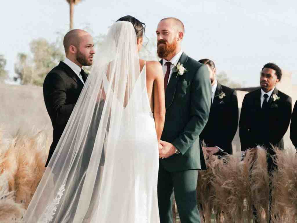 Noivos casando, com padrinhos no fundo, estão ao ar livre