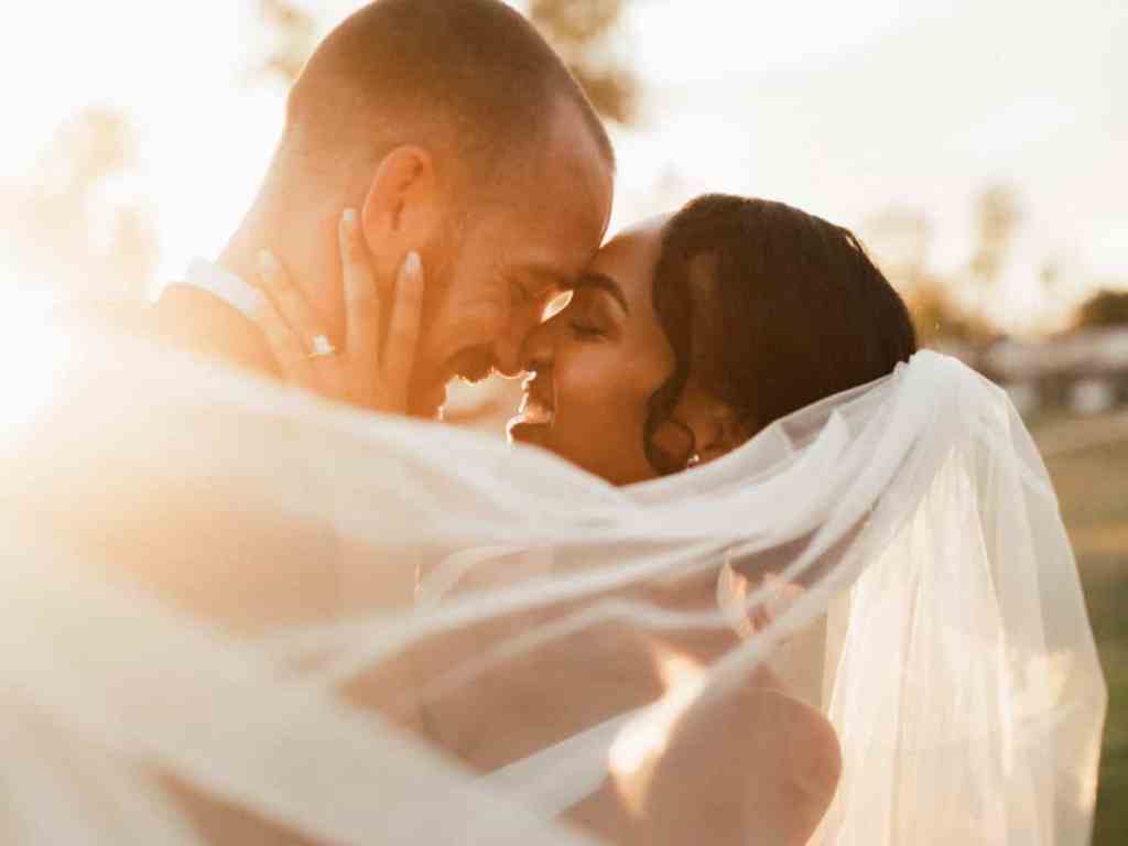 Noivos com a testa encostadas e sorrindo, o véu dela está na parte da frente voando