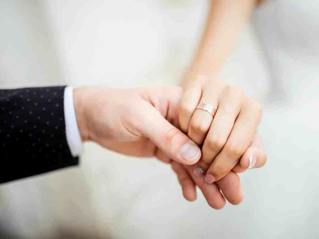 Noivos segurando a mão um do outro, na dela é possível ver uma aliança larga