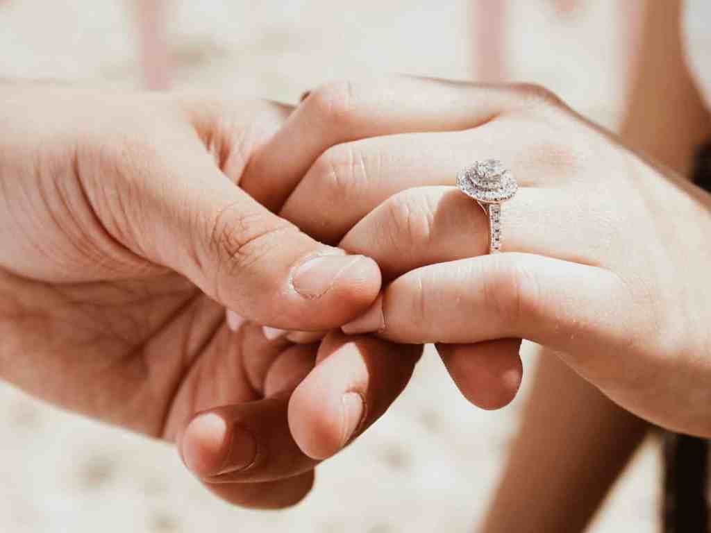 Homem segurando mão da noiva, que usa anel de noivado com pedra central grande - como acertar na aliança?