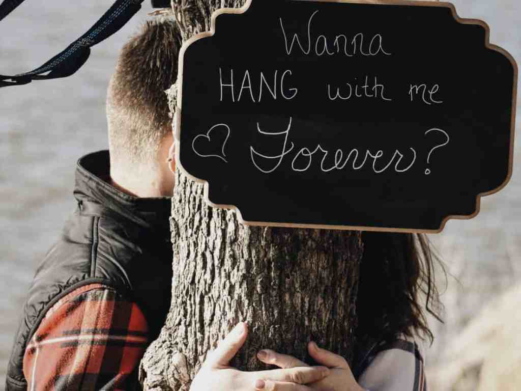 Placa amarrada na árvore com a frase em inglês "quer sair comigo para sempre? ", de fundo uma praia