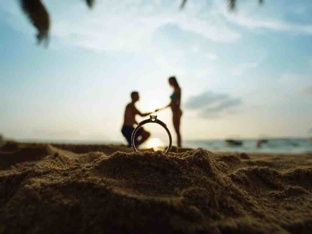 Homem ajoelhado fazendo pedido de casamento na praia, enquanto na frente, na areia há um anel de noivado