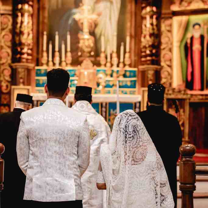 Quanto custa um casamento no Brasil e no mundo? - Casamento-na-igreja-catolica Casamento na igreja católica, noivos estão de costas para a foto e ao fundo os detalhes da igreja católica desfocados como velas, imagens.