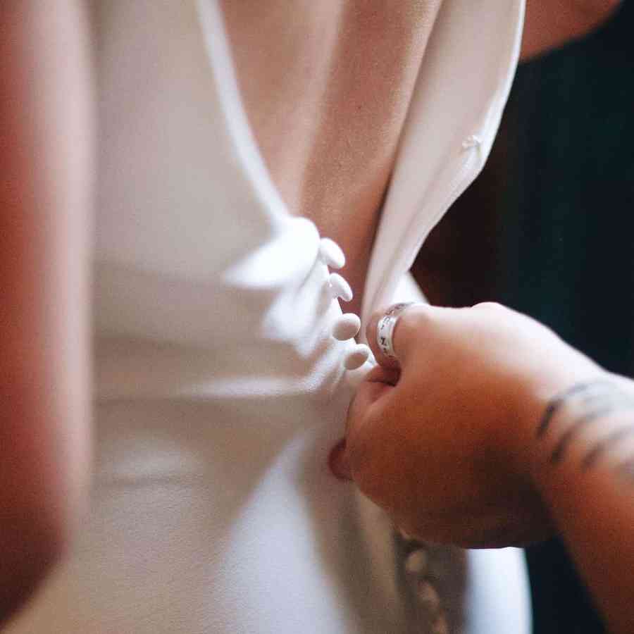 Imagem de uma noiva se arrumando para o casamento, está fechando os botões do vestido de noiva atrás.