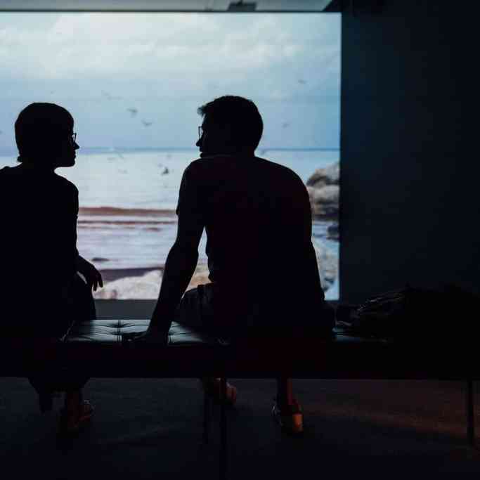Casal conversando em frente a um refletor de imagens que mostra a praia.