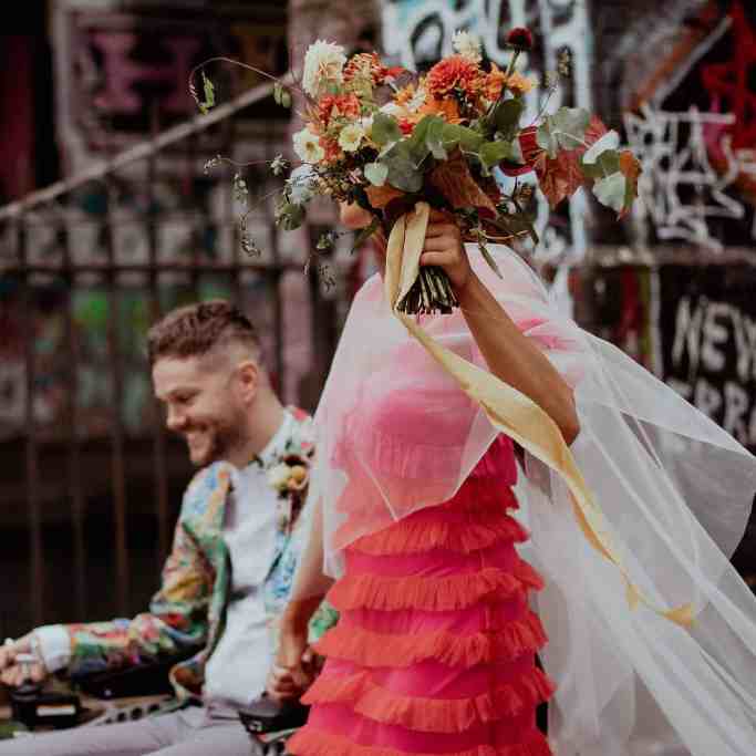 Noiva de vestido rosa e buquê, enquanto segura mão do noivo de cadeiras de rodas
