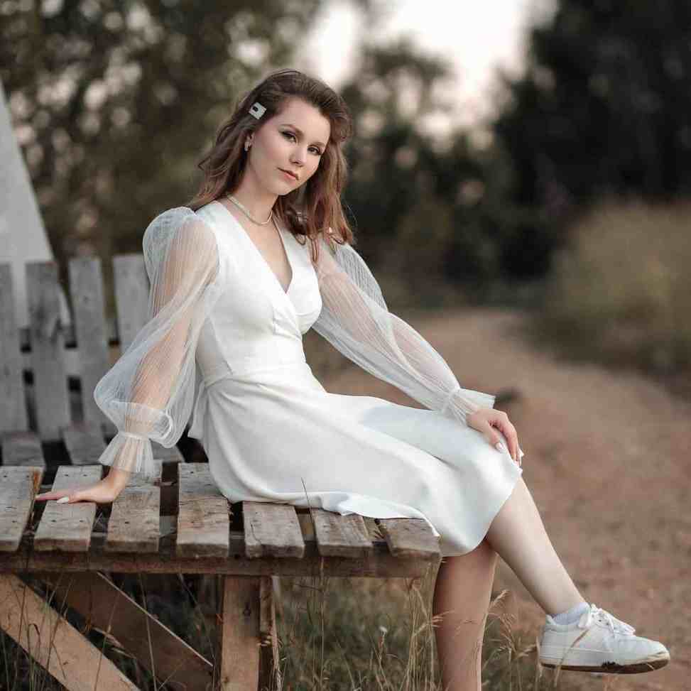Mulher de vestido branco e tênis branco sentada em um banco de madeira