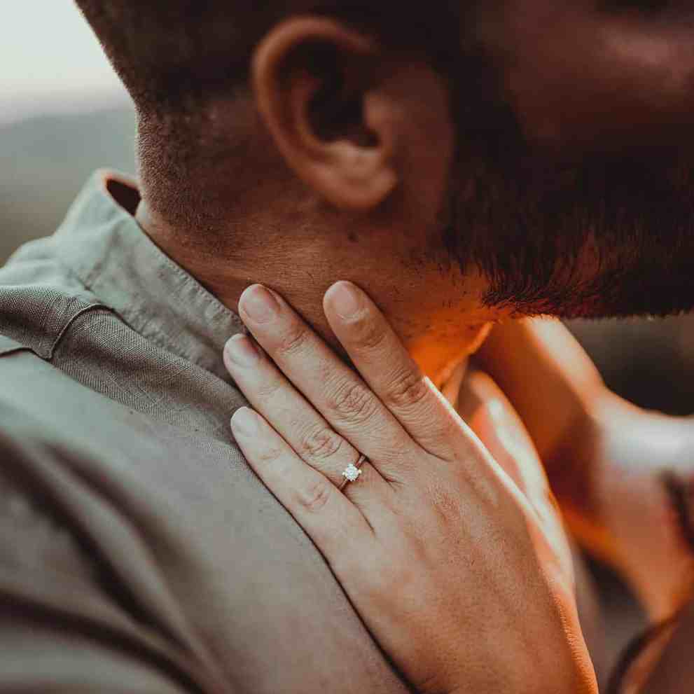 Casal se beijando, o foco da foto é na mão dela com anel de noivado