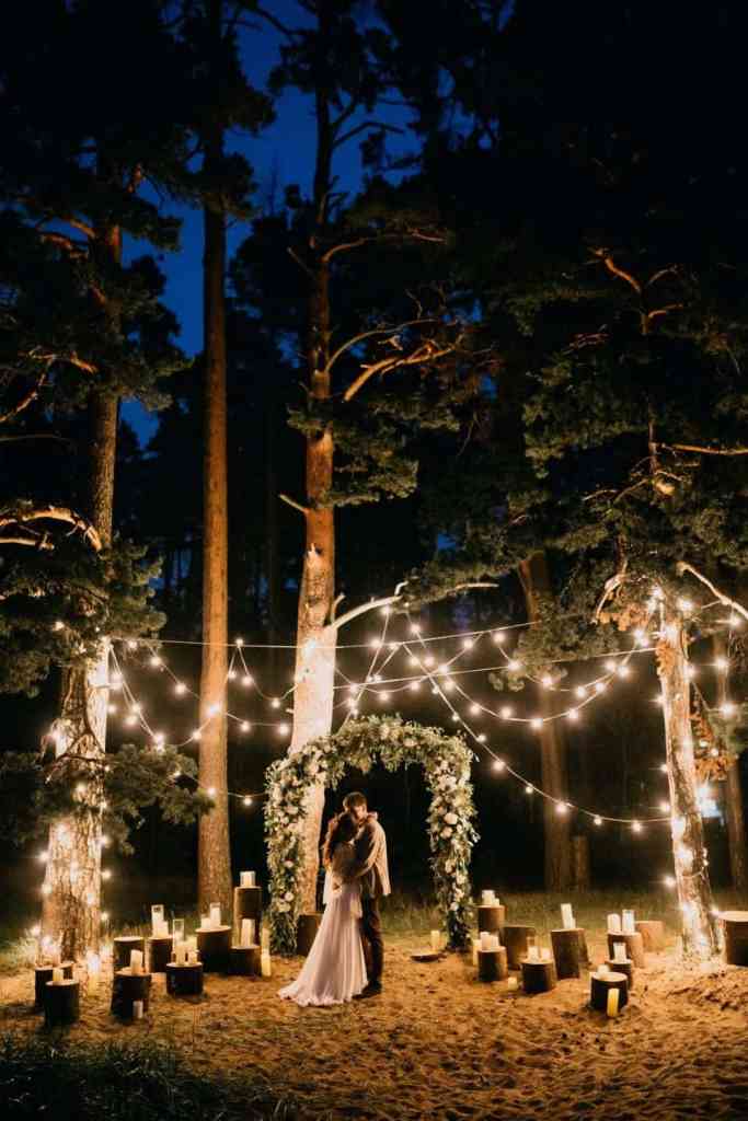 Noivos abraçados em casamento ao ar livre, com decoração cheia de luzes de velas e de lâmpadas