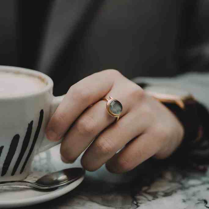 Pessoa tomando café, no dedo do meio possui um anel de ouro com pedra verde