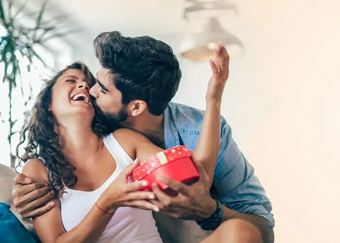 Homem beijando namorada na bochecha enquanto ela ri e recebe um presente