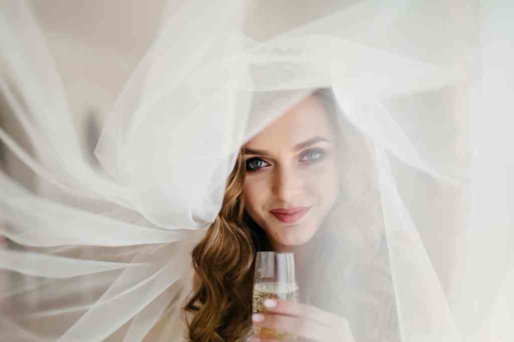 Noiva segurando taça de champagne e seu véu está cobrindo ao redor da câmera