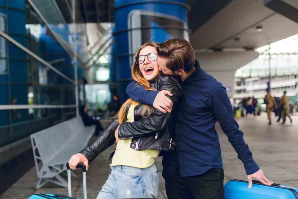 Homem e mulher abraçados, segurando malas de viagem em frente ao aeroporto
