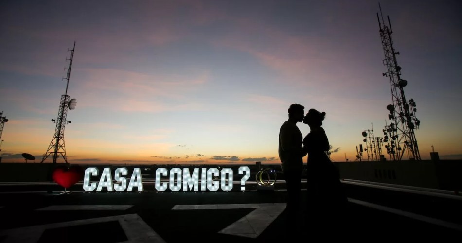 Foto com penumbra, é possível ver a sombra de um casal abraçado e o letreiro escrito casa comigo brilhante no chão